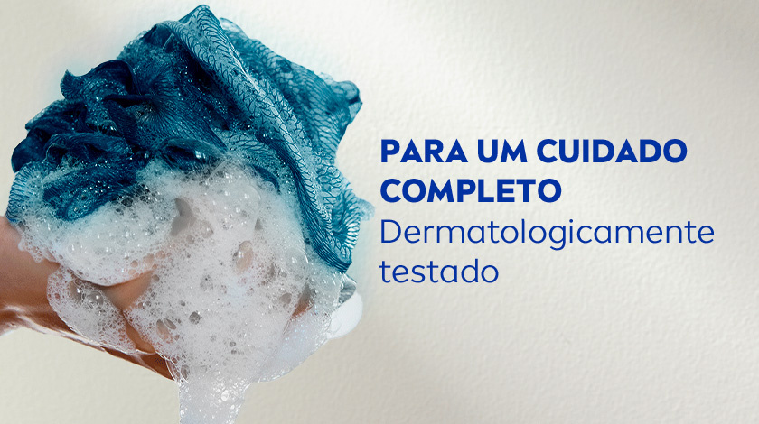Close de uma mão segurando uma bucha azul cheia de espuma branca em um fundo neutro.