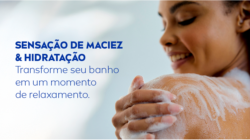 Mulher sorridente tomando banho, aplicando espuma branca no ombro com a mão direita. Fundo desfocado em tons azulados.