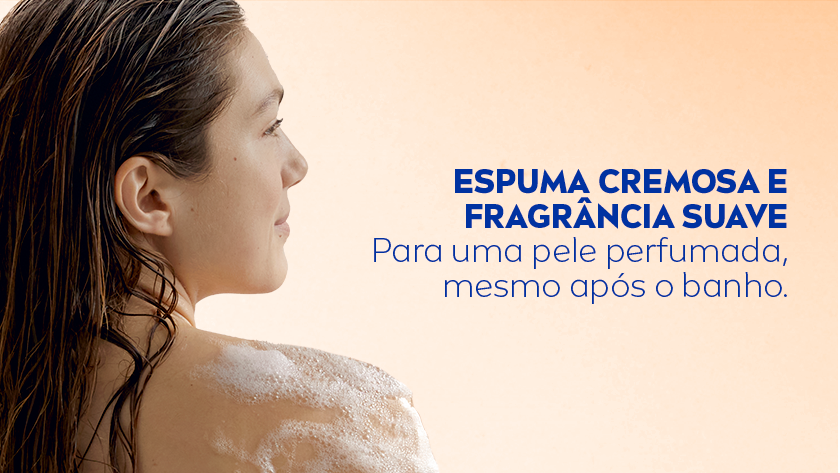 ESPUMA CREMOSA E FRAGRÂNCIA SUAVE Para uma pele perfumada, mesmo após o banho. Mulher de perfil, com cabelos castanhos longos e molhados, aparece de costas durante o banho. Há espuma visível sobre seu ombro, sugerindo o uso de sabonete. O fundo é cor de salmão. À direita da imagem, em letras azuis, está o texto.