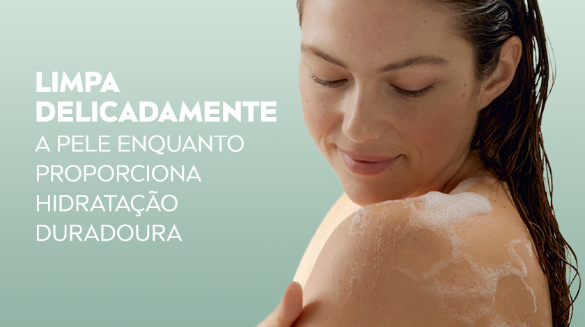 Mulher de cabelos escuros tomando banho, ensaboando os ombros, com expressão relaxada, sobre fundo verde claro.