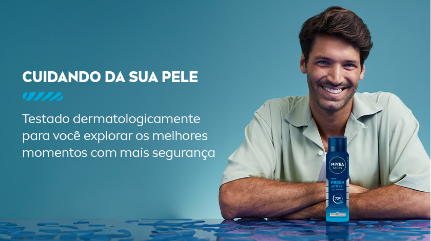 Um homem com os braços apoiados em uma mesa azul sorri, ele usa uma camisa verde clara. Em sua frente, é posicionado o desodorante aerossol. O fundo é azul claro.
