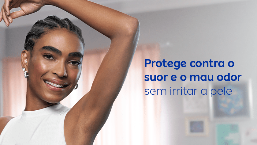 Uma mulher sorri, enquanto levanta o braço e exibe sua axila. Ela veste uma regata branca, e o ambiente é neutro.