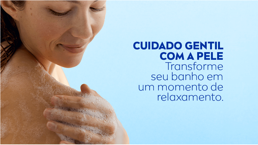 Mulher com expressão relaxada ensaboa o ombro com espuma abundante durante o banho, em um fundo azul claro.