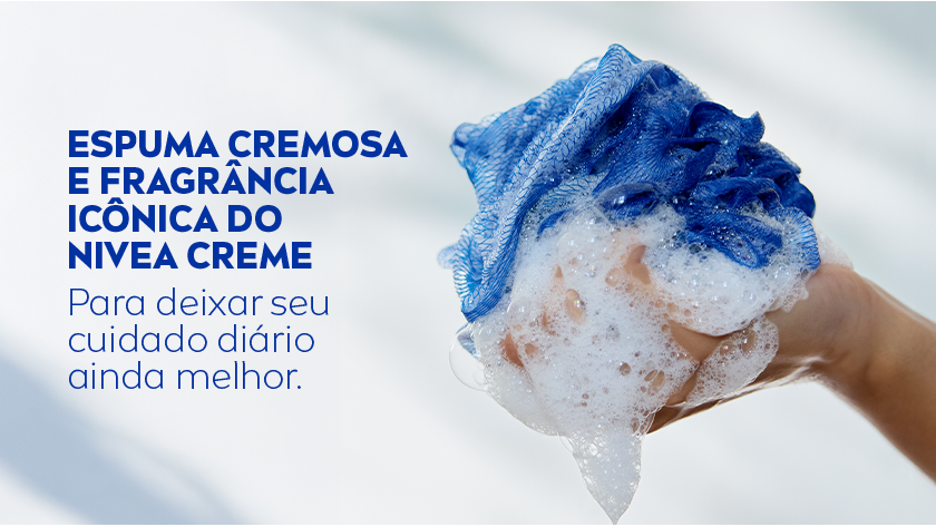 Mão segurando uma esponja de banho azul com espuma branca. Fundo claro e suave.
