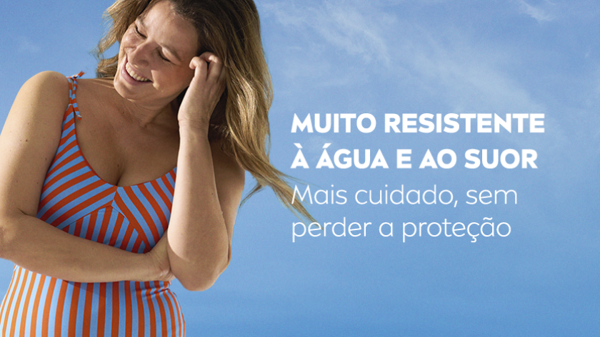 Uma mulher sorri, enquanto passa a mão na cabeça, ela usa um maiô de praia. O fundo é um céu azul.