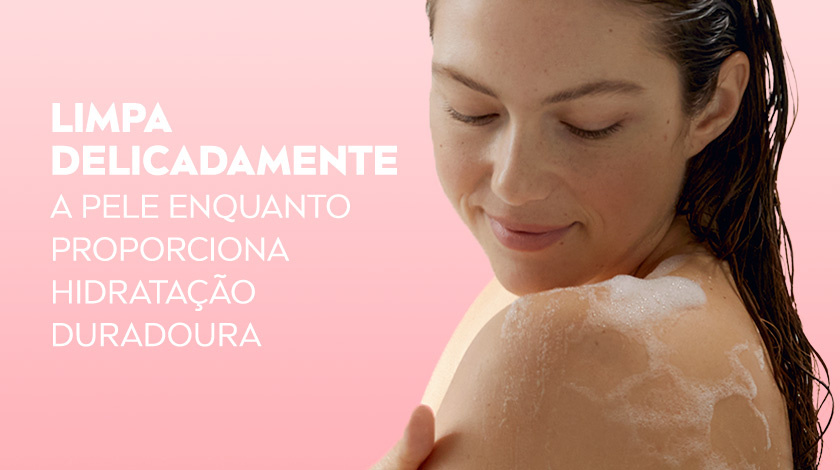 Mulher de cabelos escuros tomando banho, sorrindo levemente enquanto ensaboa o ombro, com espuma sobre a pele.