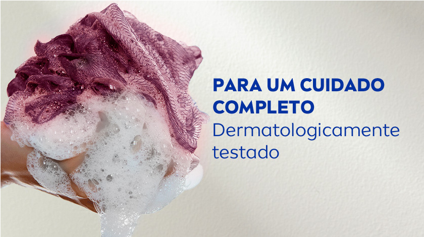 Para um cuidado completo dermatologicamente testado Close de uma mão segurando uma bucha roxa cheia de espuma branca em um fundo neutro.