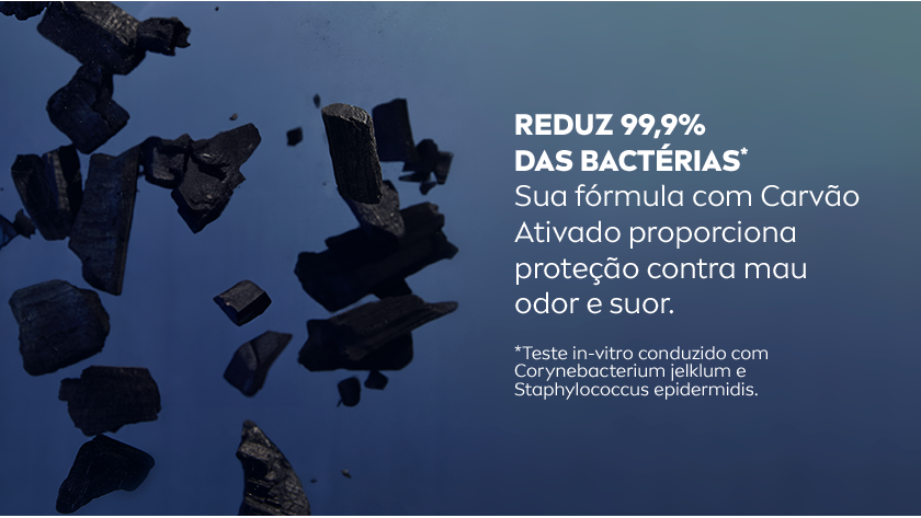 Fundo azul escuro e gradiente para azul claro. Fragmentos de carvão ativado aparecem flutuando em primeiro plano, simbolizando a presença do ingrediente na fórmula. À direita, texto em letras brancas destaca: “REDUZ 99,9% DAS BACTÉRIAS*” seguido da descrição dos benefícios do carvão ativado, com letras menores em tom branco. Pequena observação em letras brancas na parte inferior informa o teste in-vitro realizado com bactérias.